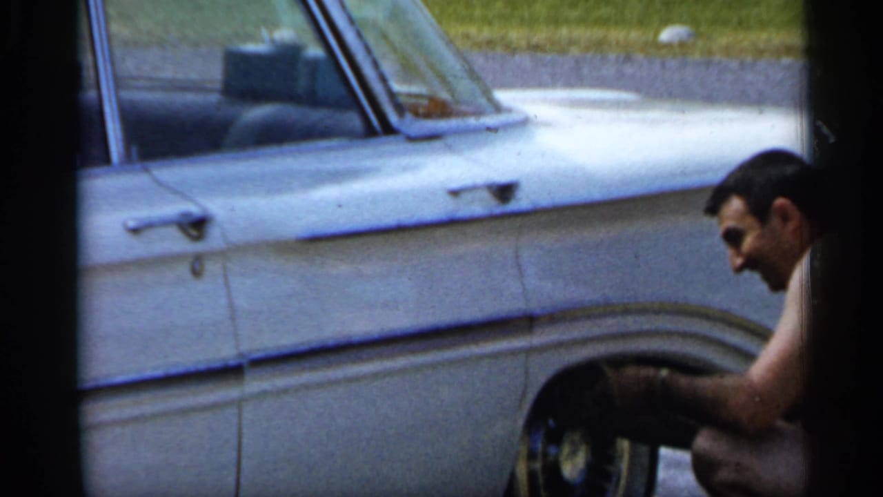 Shirtless man crouches beside the front wheel of a white classic car.