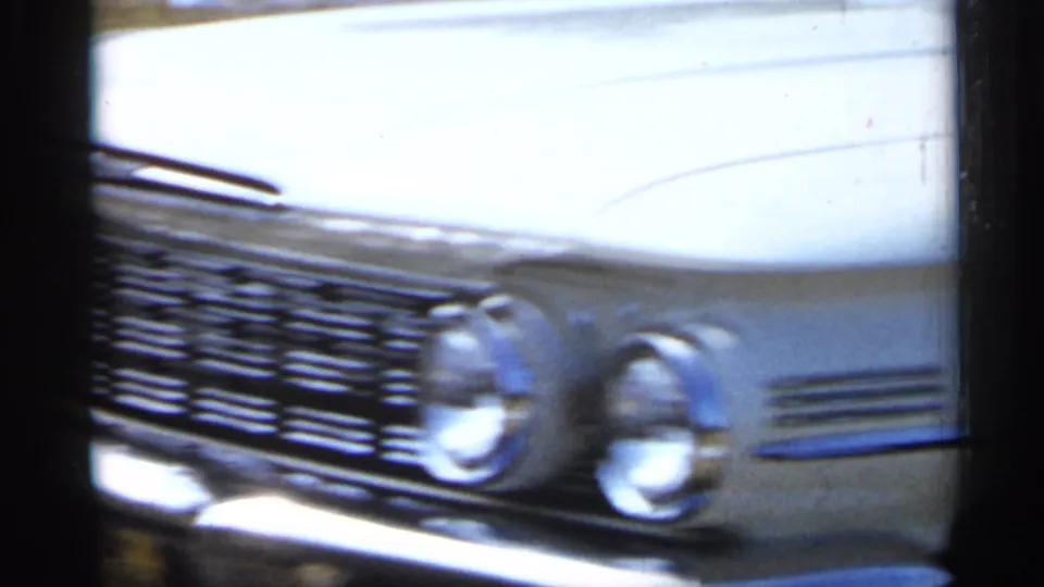 The camera moves close across the white classic car's front end, showing the chrome grille, hood, and round headlights in soft 8mm color.