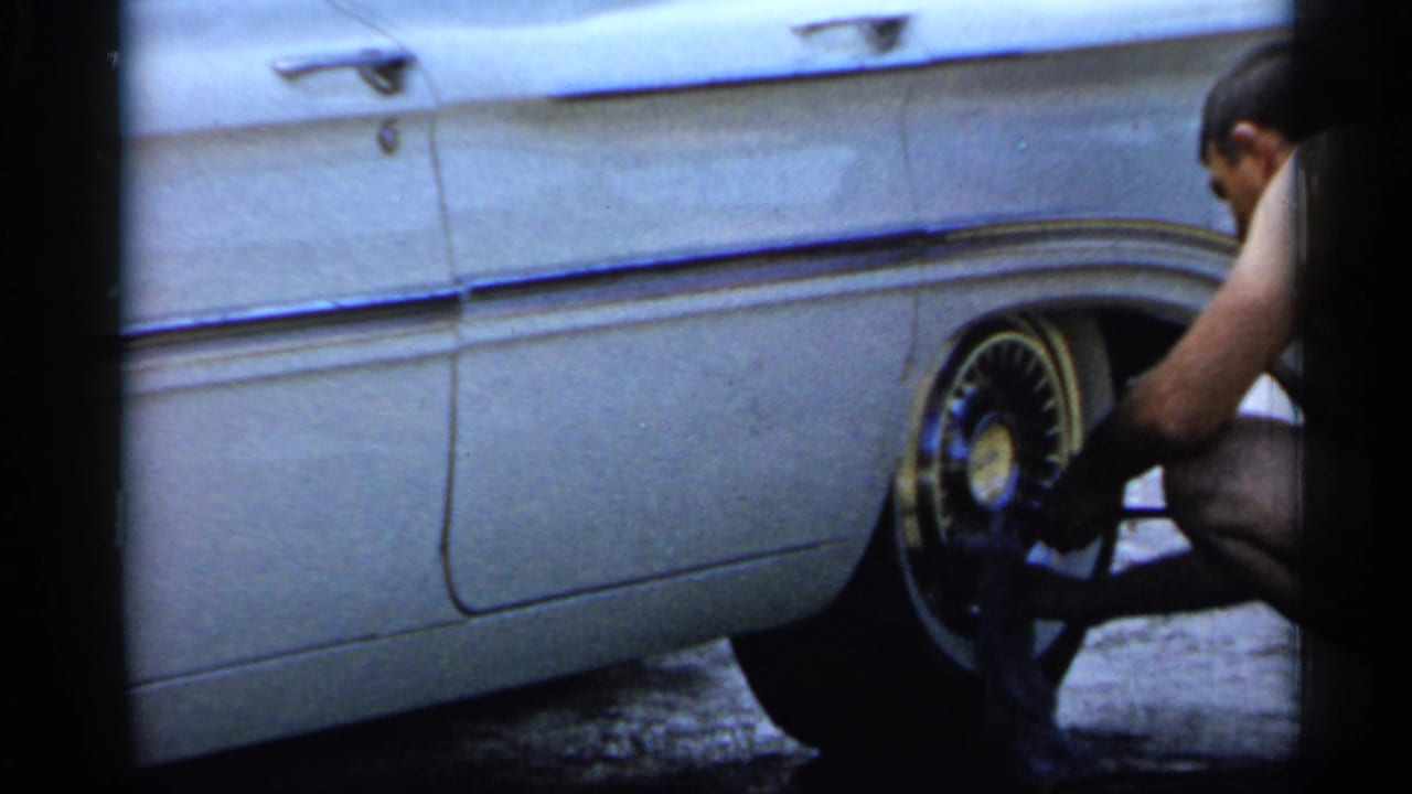 1958 Florida home-movie frame of a shirtless man crouched by the rear wheel of a light-colored car, handling the tire or hubcap.