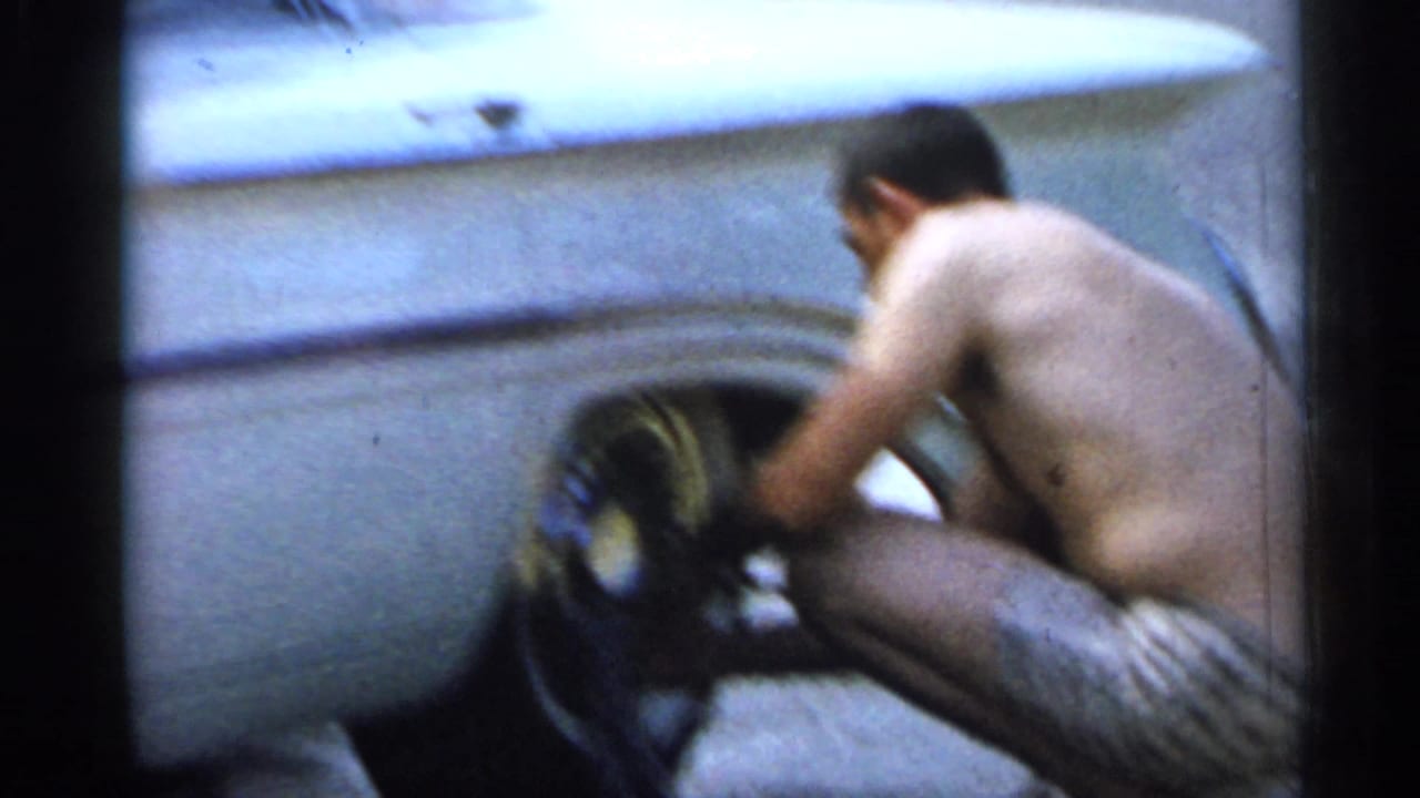 A shirtless man in swim trunks works at the wheel of a light-colored car in a soft, grainy 8mm Florida vacation scene.