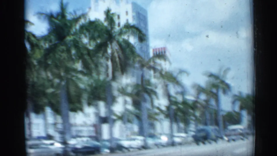 Passenger-view 8mm frame from a moving vehicle passing palm trees, traffic, and tall Florida buildings in 1958.
