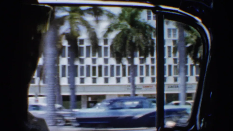 Inside a moving car in 1958 Florida, silhouetted occupants face a window with palm trees, urban buildings, and vintage traffic outside.
