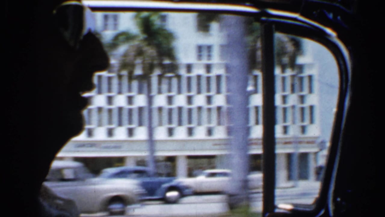 A dark passenger profile is seen inside a moving car, with 1958 Florida street traffic, palm trunks, and a large building beyond the window.