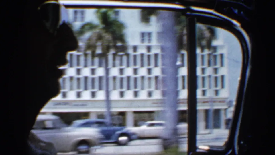A dark passenger profile is seen inside a moving car, with 1958 Florida street traffic, palm trunks, and a large building beyond the window.