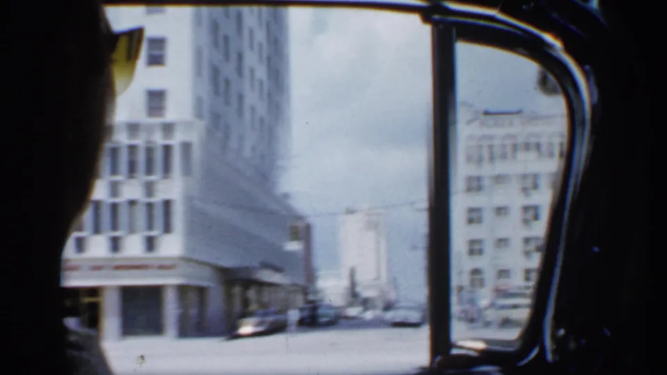 From inside the car, the camera looks toward a Florida street corner with tall buildings, palm trees, and a dark occupant at the window edge.