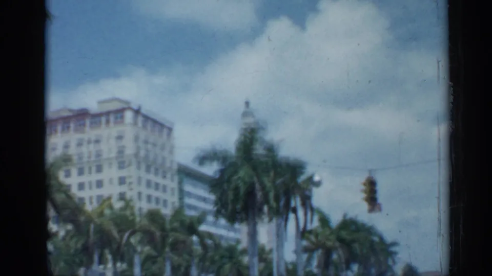 The moving car view shows a palm-lined Florida city block with multi-story buildings, a traffic signal, clouds, and soft 8mm color.