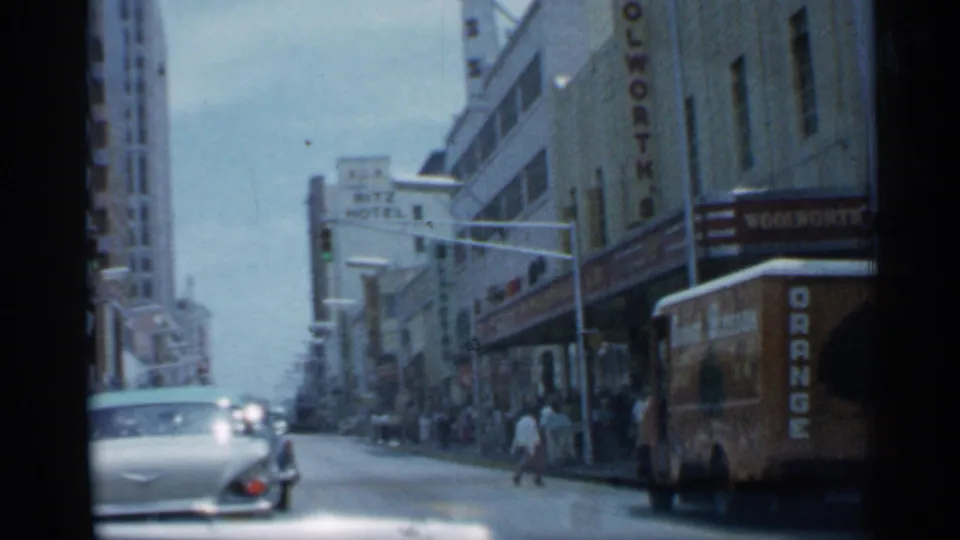 1958 Florida downtown street with classic cars, pedestrians, storefronts, a visible Woolworth's sign, and a large vehicle along the curb.