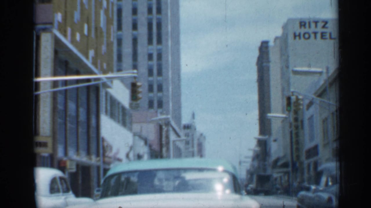 1958 Florida street view from inside a car, with a pale automobile ahead, tall buildings, traffic signal, and Ritz Hotel sign visible.