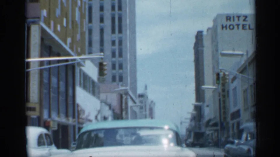 1958 Florida street view from inside a car, with a pale automobile ahead, tall buildings, traffic signal, and Ritz Hotel sign visible.