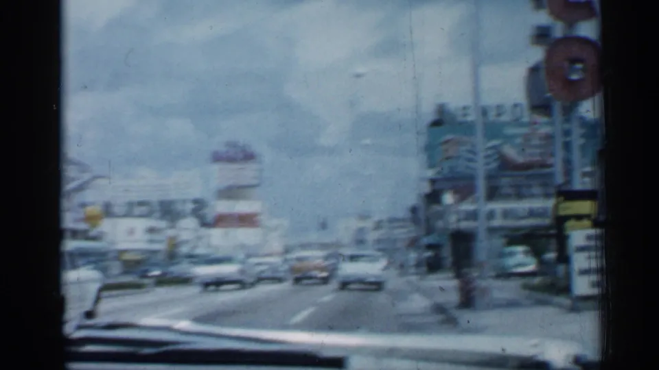 1958 Florida home-movie frame from inside a moving car, showing a multi-lane commercial street with automobiles, signs, and storefronts.