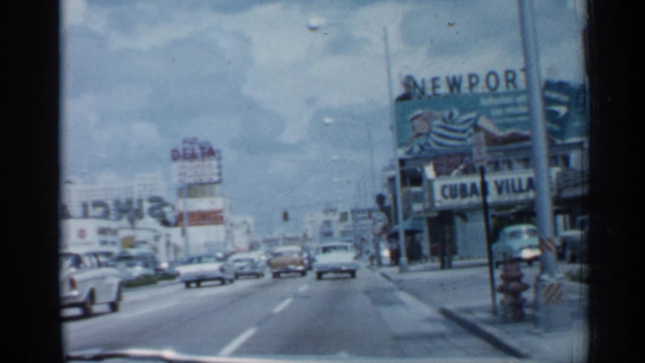 Moving street view with classic cars, Newport and Cuban Village signs, and commercial buildings.