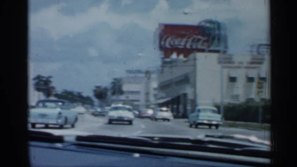 1958 Florida driving frame showing classic cars, palm-lined roadside space, and a large Coca-Cola sign on a commercial building.