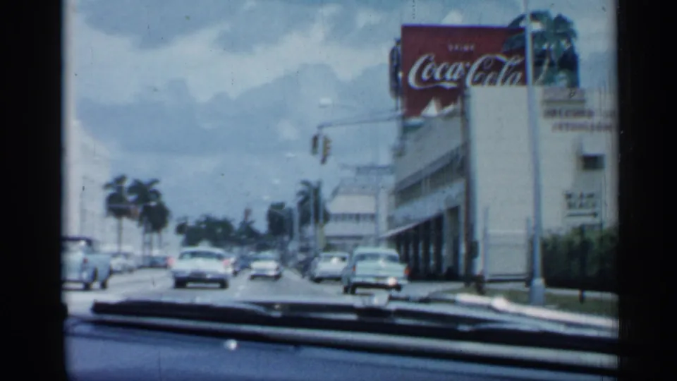 1958 Florida home-movie frame from inside a car, passing a commercial building with a Coca-Cola sign and traffic ahead.