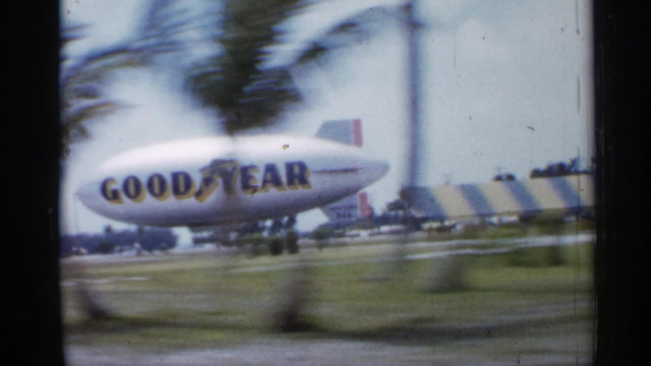 Goodyear blimp seen through a moving car window with palm trunks in the foreground.