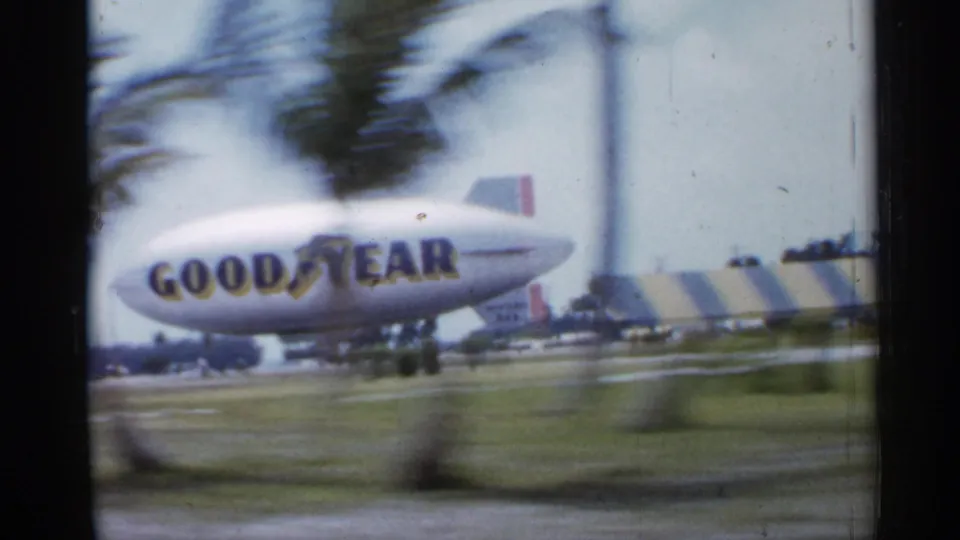 1958 Florida 8mm frame showing a Goodyear blimp beyond a roadside, partially obscured by blurred palm trunks.