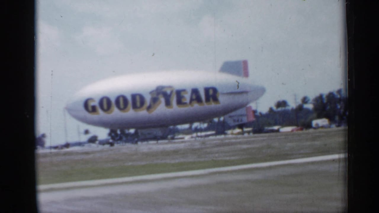 Goodyear blimp over an open roadside field with low buildings in the distance.