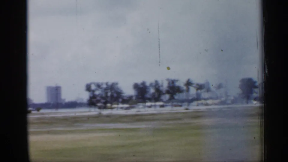 1958 Florida 8mm frame showing a hazy roadside view with grass, trees, low buildings, and a distant vertical building.