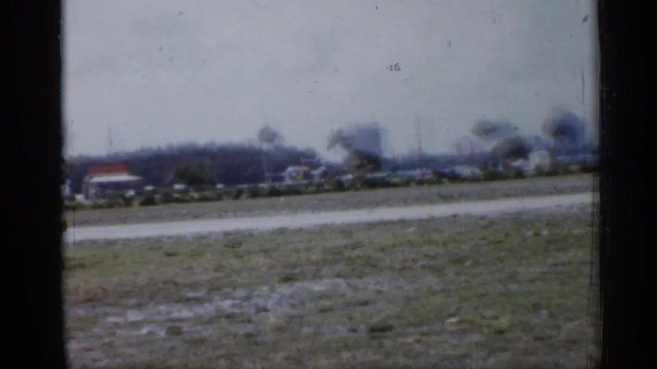 1958 Florida home-movie frame showing a soft, distant roadside landscape with grass, trees, road, and low buildings.