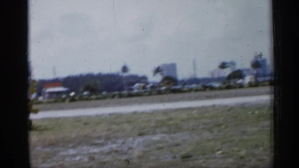 1958 Florida home-movie frame with open grass, a low road or waterfront edge, distant palms, and soft buildings beyond a dark window-like border.
