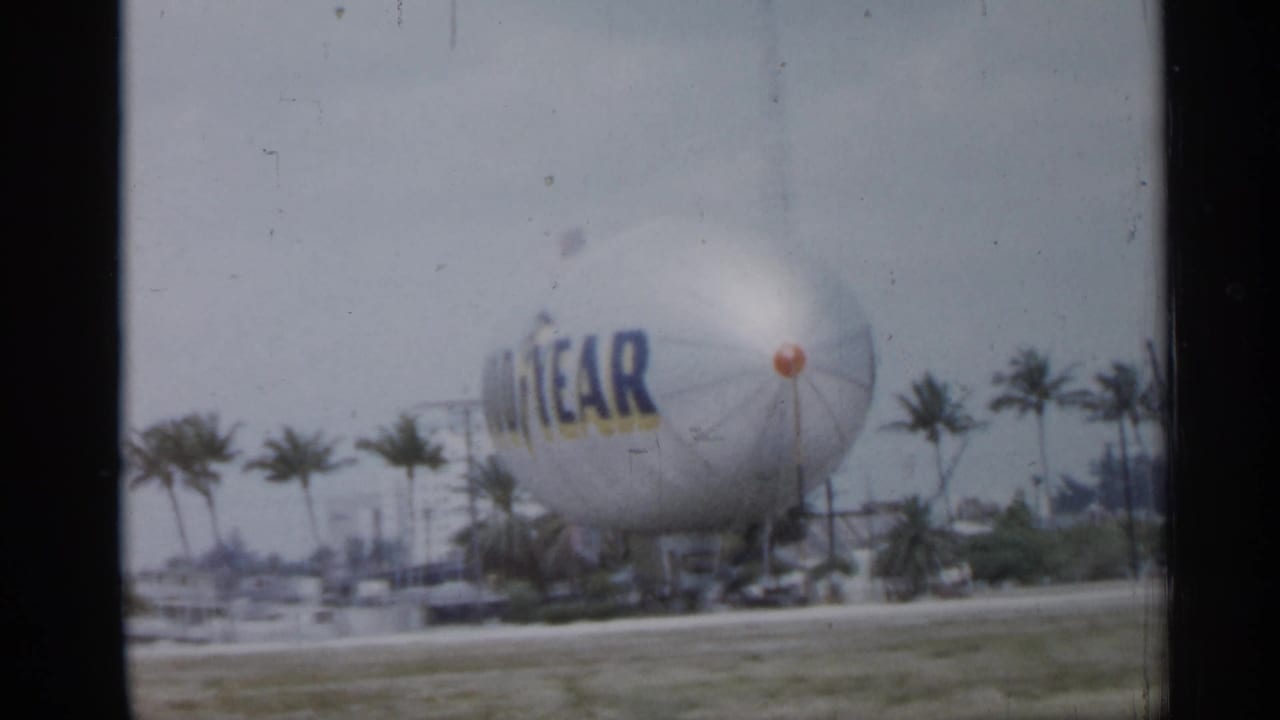 1958 Florida home-movie frame showing a Goodyear blimp grounded in a grassy area beyond a roadside with palm trees nearby.
