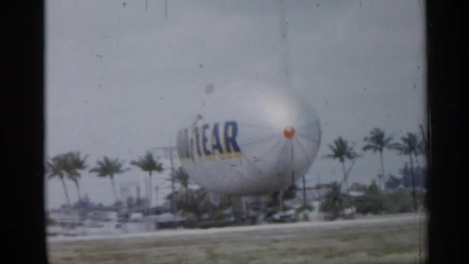 1958 Florida home-movie frame showing a Goodyear blimp grounded in a grassy area beyond a roadside with palm trees nearby.