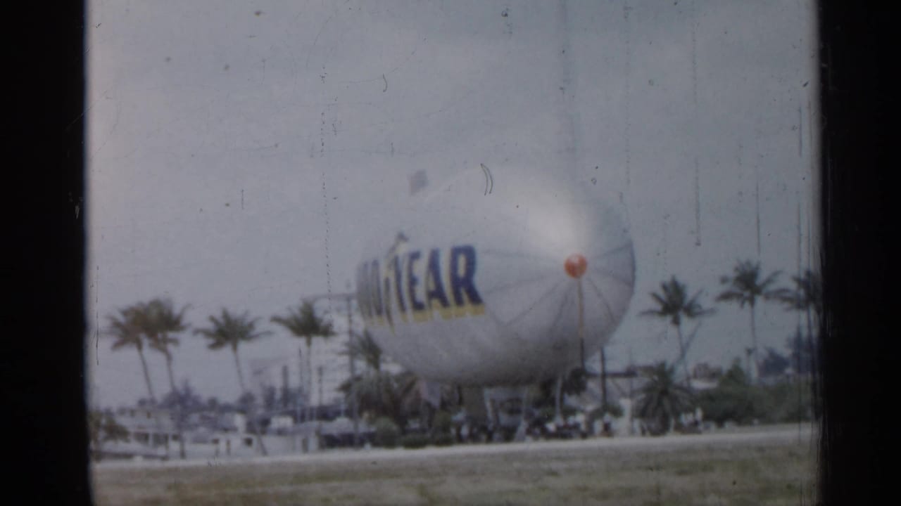The Goodyear blimp appears grounded beyond a roadway in Florida, with palm trees and soft overcast light in the frame.