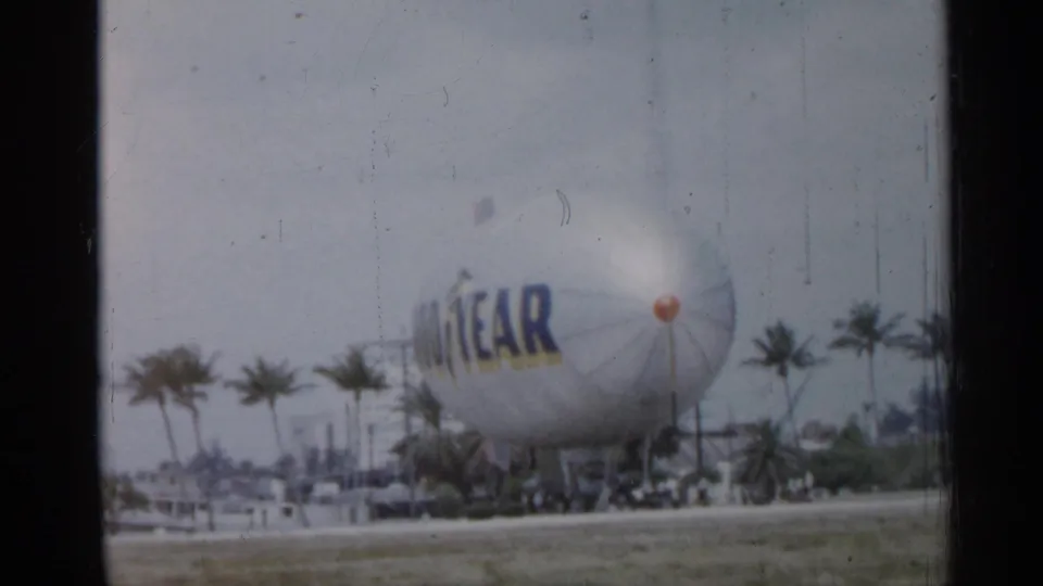 The Goodyear blimp appears grounded beyond a roadway in Florida, with palm trees and soft overcast light in the frame.