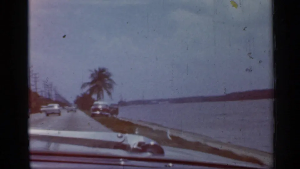 1958 Florida home-movie frame from a moving car, showing a waterfront road, parked or passing cars, palms, and open water.