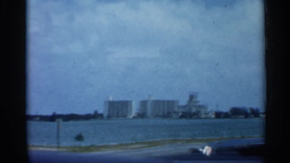 1958 Florida home-movie view across water toward distant mid-century high-rise buildings, partly framed by a dark car-window edge.