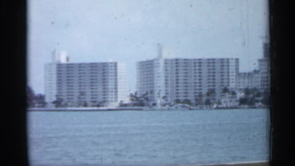 Florida waterfront scene with pale mid-century high-rise buildings across calm water under a hazy blue sky.