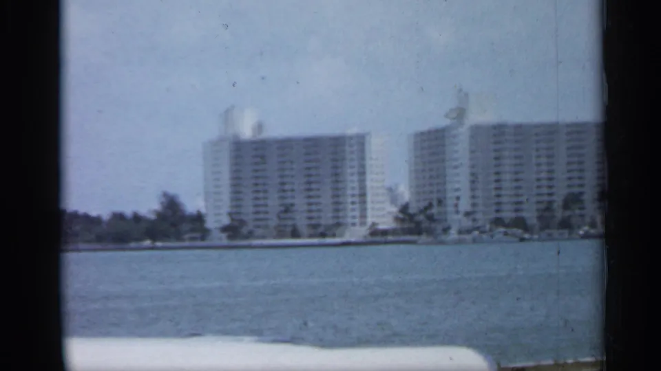 1958 Florida shoreline with tall pale buildings, trees, and open water, captured in soft 8mm color.