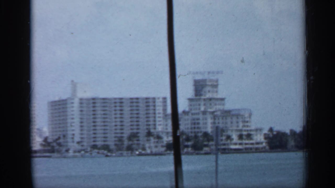 1958 Florida waterfront view with white high-rise buildings across the water, framed by dark film edges and a vertical obstruction.