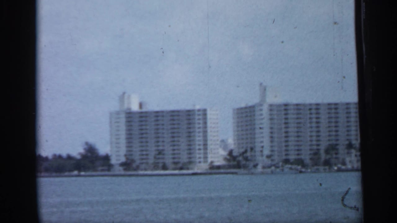 1958 Florida coastal view showing modern waterfront towers across bay water, with shoreline trees and soft 8mm film grain.