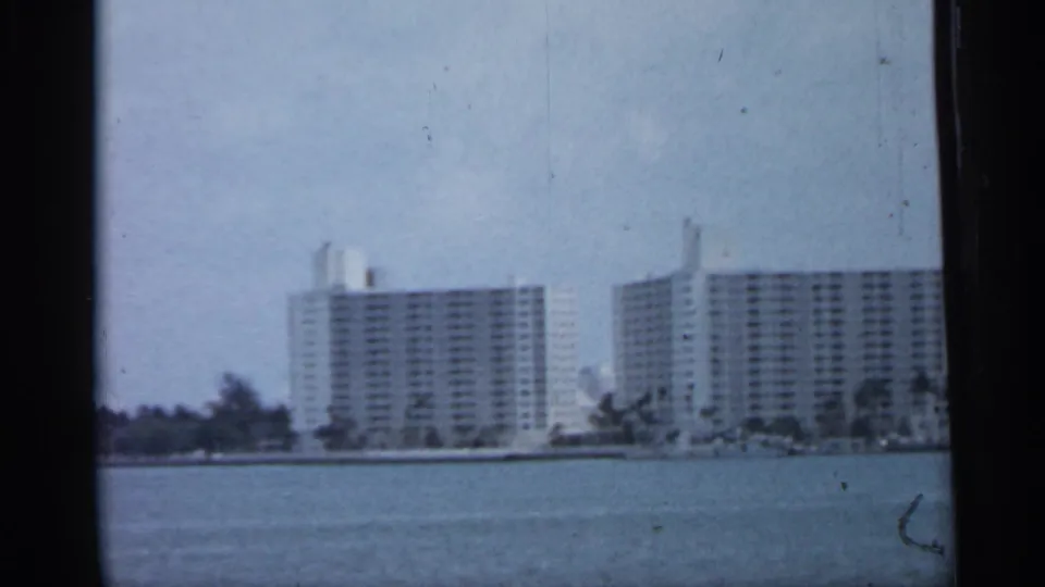 1958 Florida coastal view showing modern waterfront towers across bay water, with shoreline trees and soft 8mm film grain.