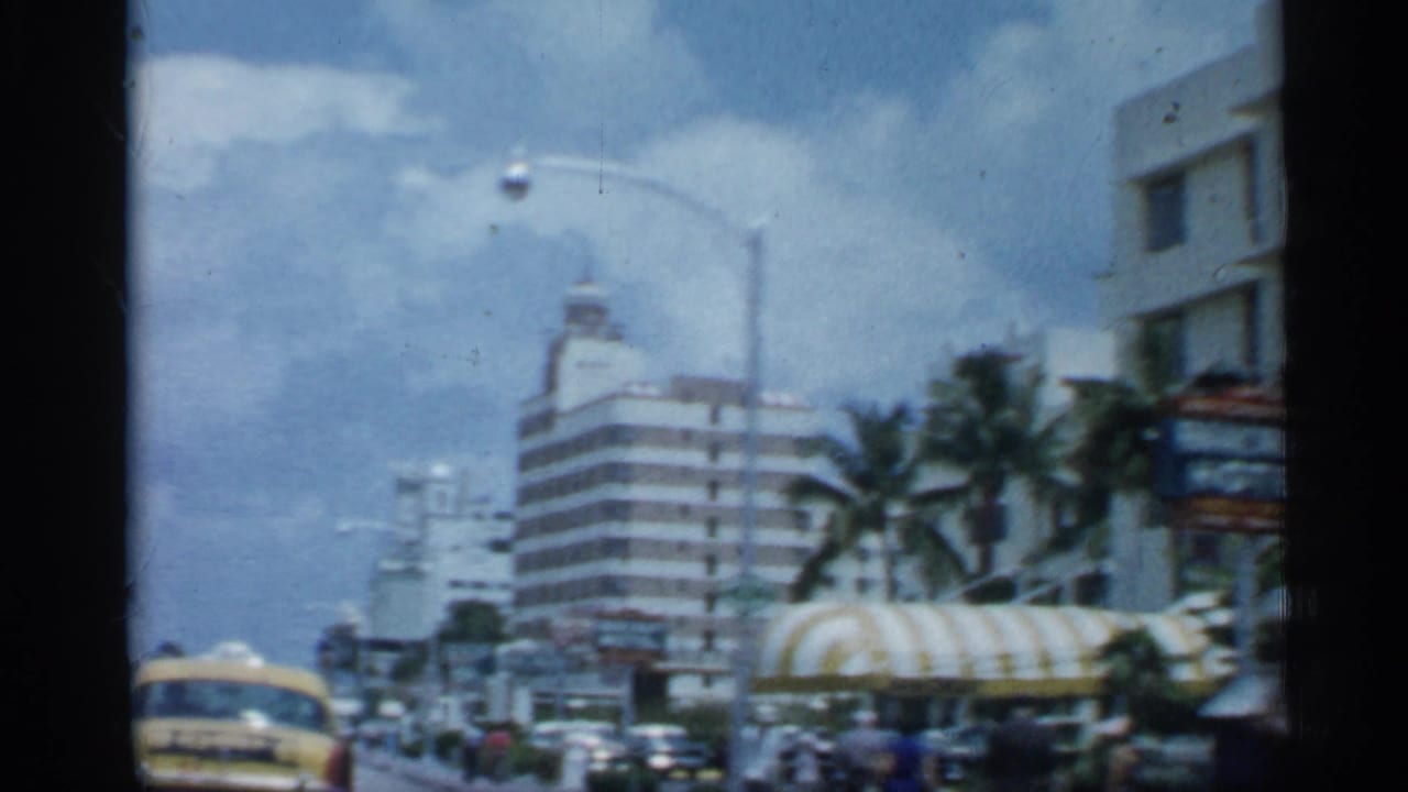 Palm-lined Florida street with mid-century cars, buildings, awnings, and a yellow taxi.