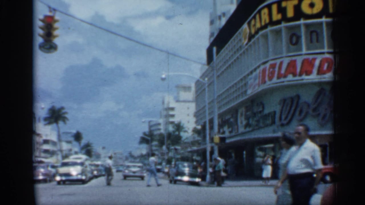 Busy street beside a Carlton theater sign with cars, palms, pedestrians, and traffic lights.