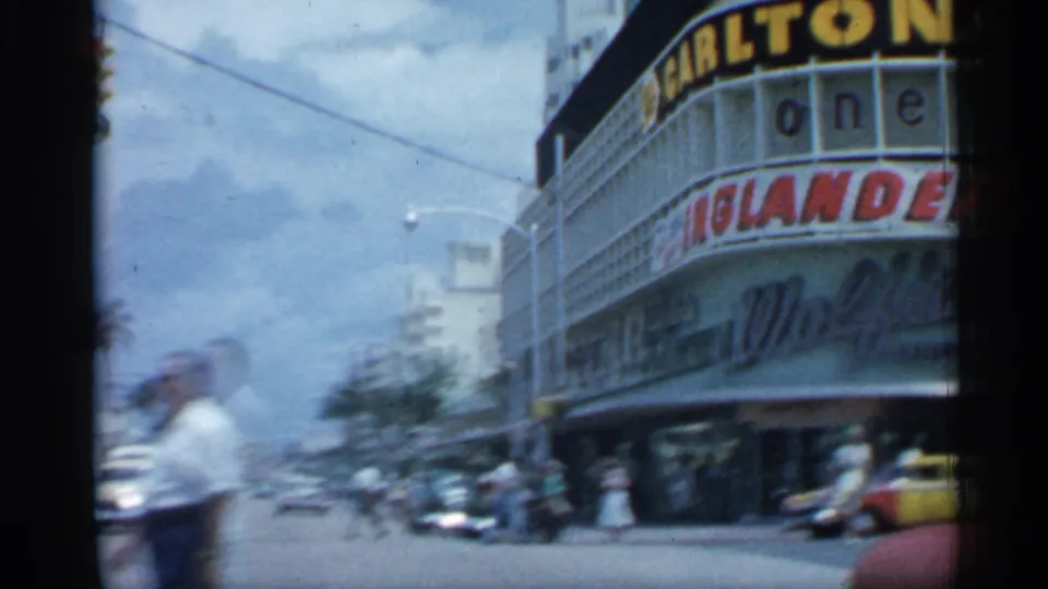 1958 Florida view of the Carlton theater corner, showing marquee lettering, passing cars, palm trees, and pedestrians along the street.