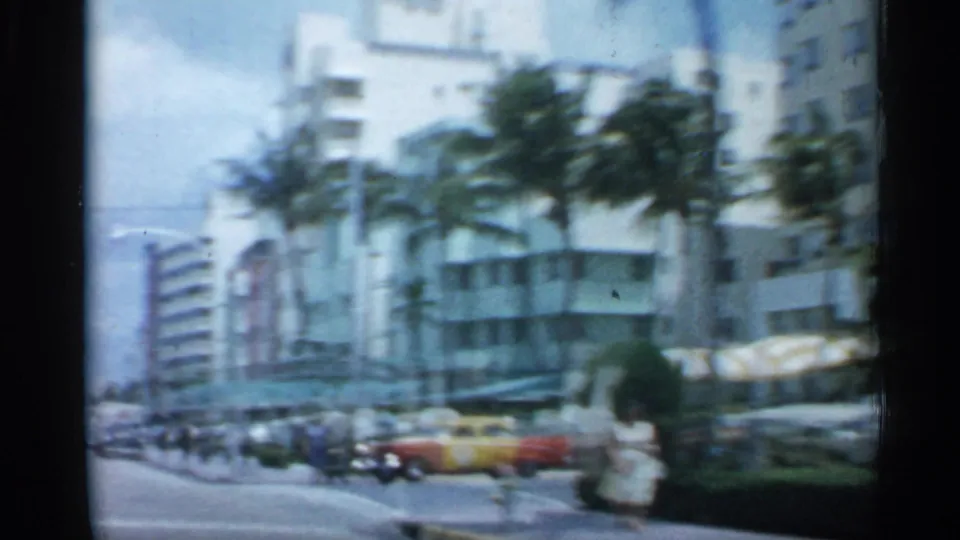 1958 Florida commercial block with palm trees, storefront awnings, pedestrians, mid-century buildings, and cars including a yellow vehicle.