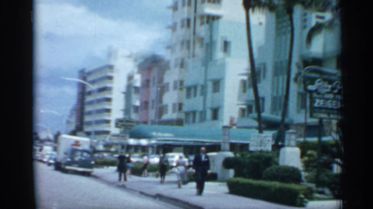 City street with pedestrians, parked cars, palm trees, awnings and business signage near tall buildings.