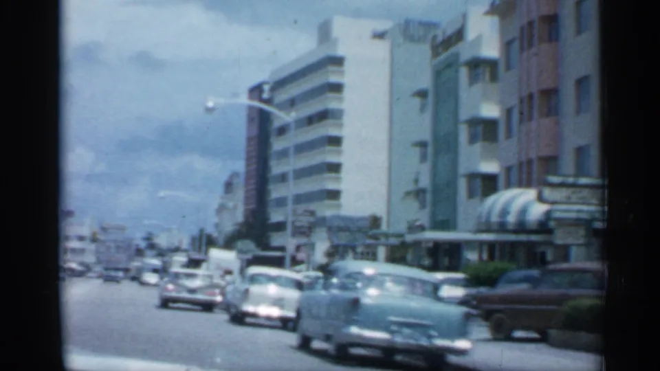 1958 Florida boulevard with classic cars, pastel building facades, striped awnings, palm trees and business signage visible from a moving camera.