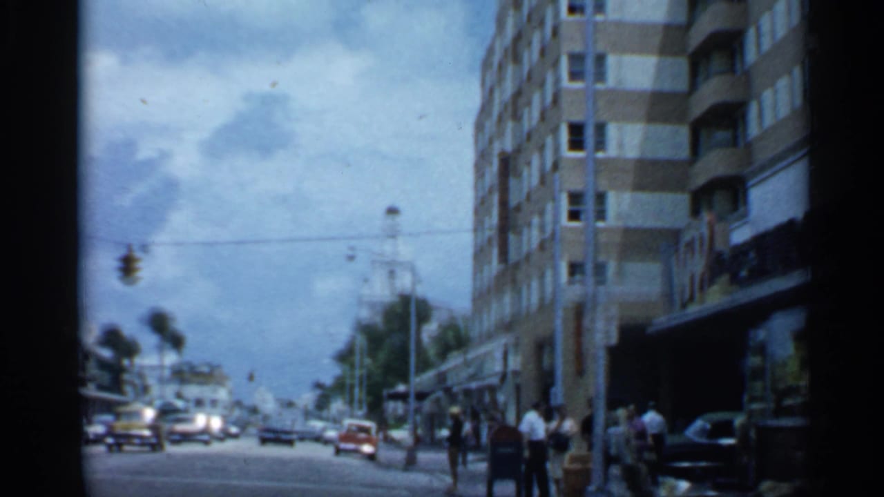 1958 Florida street view with classic cars, pedestrians near storefronts, a traffic signal, palm trees, and a tall modern building.