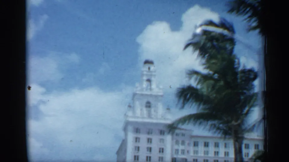 Florida street view showing a white towered building, palm fronds, clouds, and soft 8mm exposure from a moving home-movie camera.