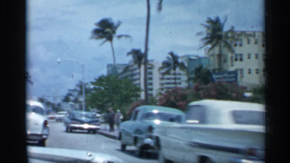 1958 Florida city street with classic cars passing, pedestrians near the roadway, palms, and multi-story buildings under a bright sky.