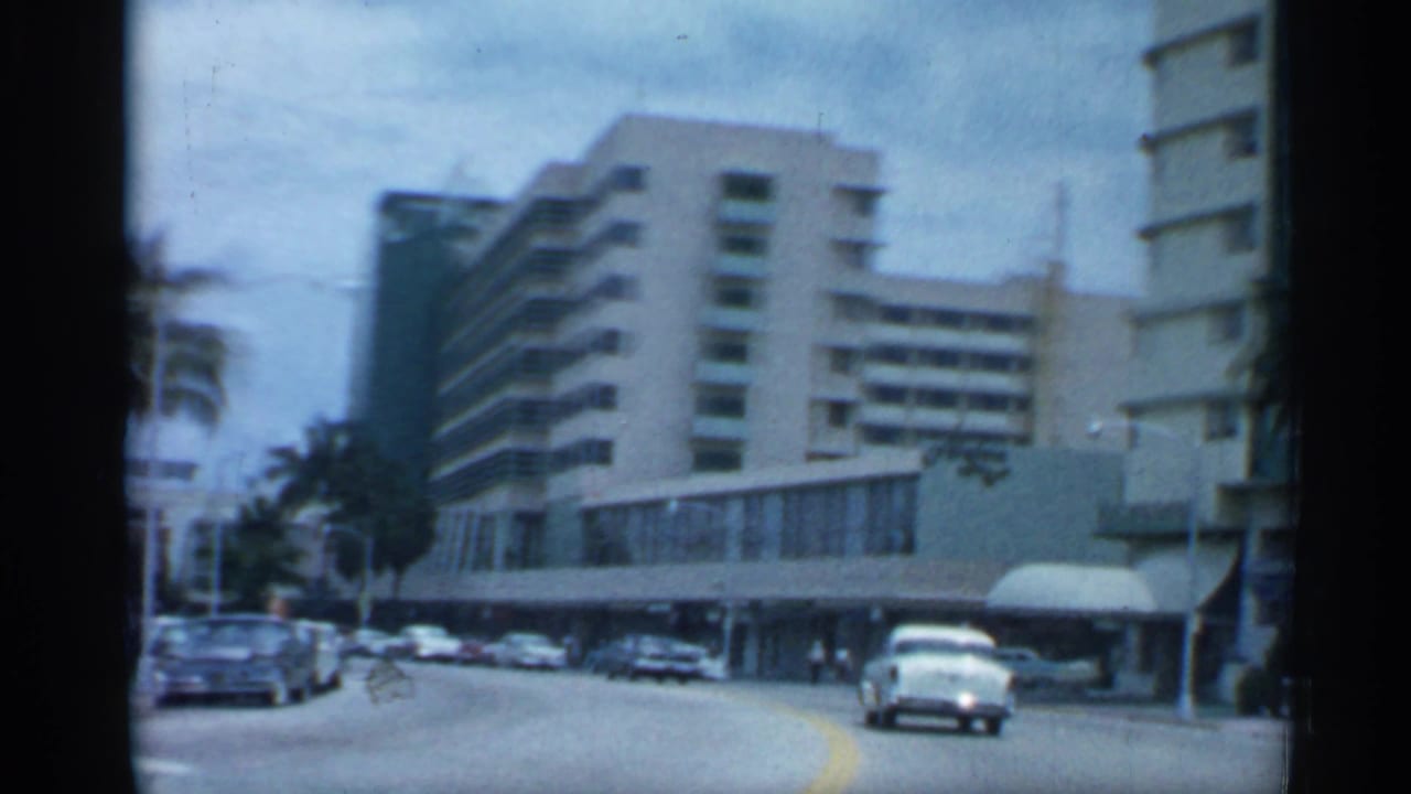 1958 Florida street view from a moving vehicle, showing classic cars, palms, and mid-century buildings along a broad urban road.