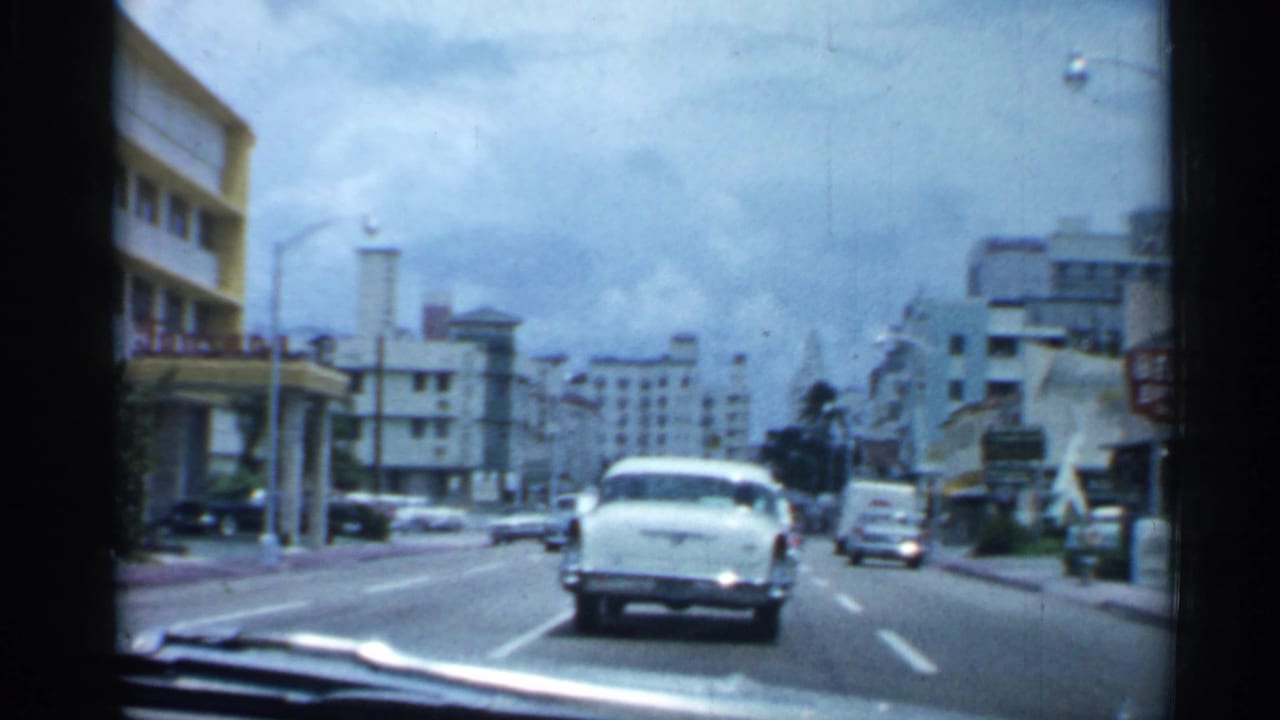 Forward view through a vehicle windshield on a 1958 Florida street, with a white car ahead and mid-century buildings on both sides.