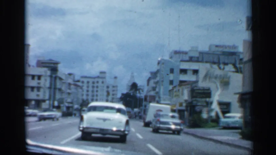 1958 Florida driving shot with a white car ahead, storefront blocks, parked vehicles, and palm trees along an urban street.
