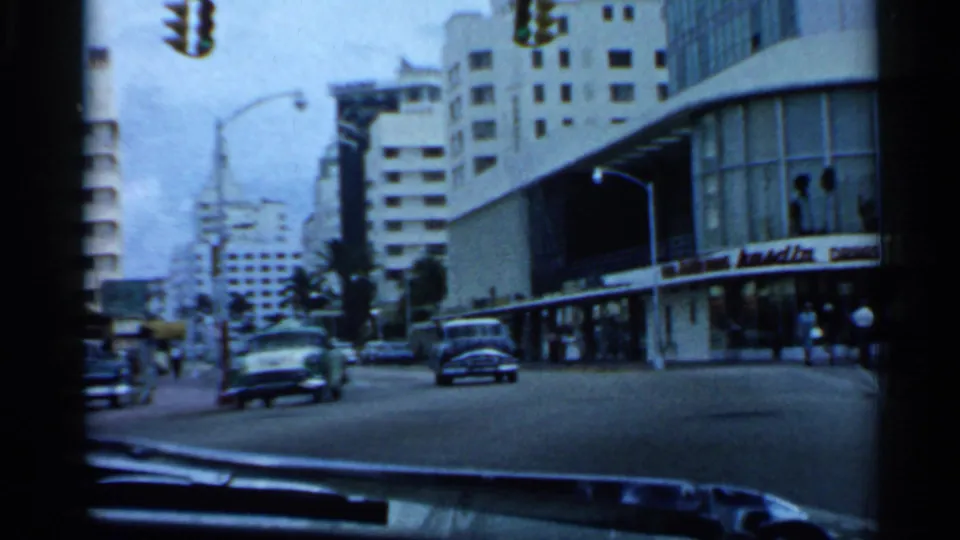 1958 Florida intersection view from a vehicle, showing traffic signals, cars, storefront signage, and a curved mid-century corner facade.