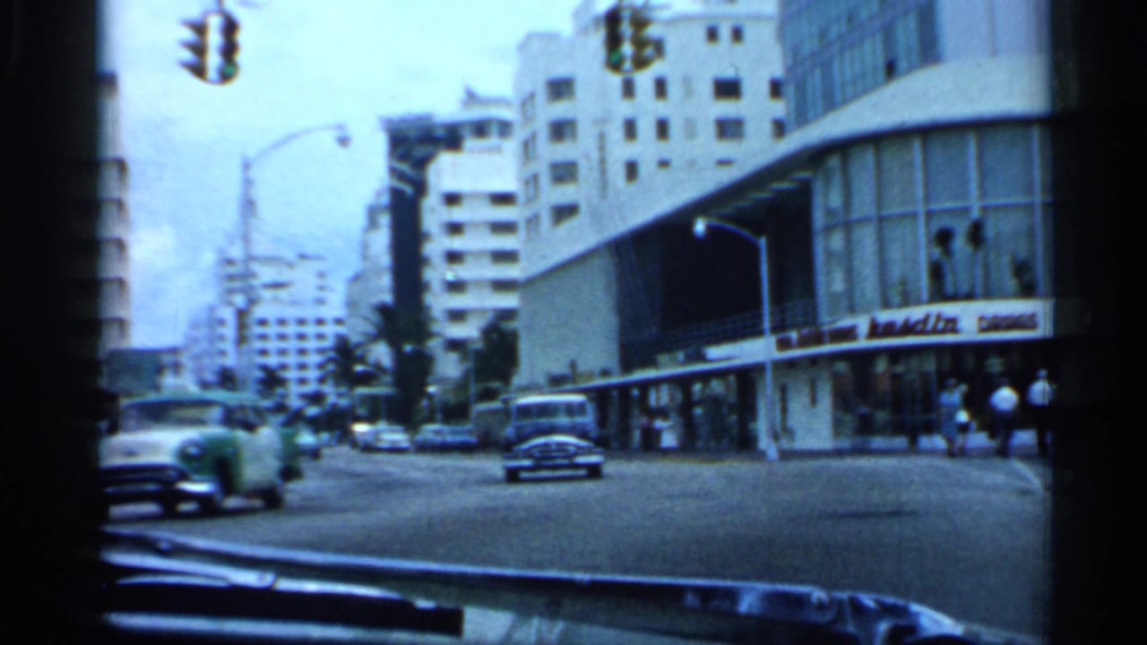 Street view from inside a car with classic automobiles, traffic lights, storefronts, and mid-century buildings.