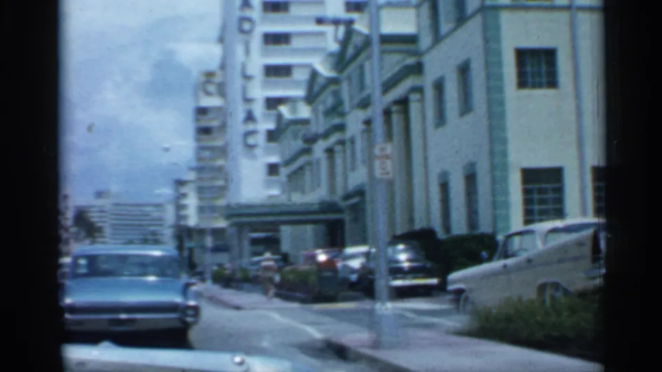 1958 Florida home-movie frame showing a classic car near the camera, a tall street fixture, parked cars, sidewalks, and mid-century urban buildings.