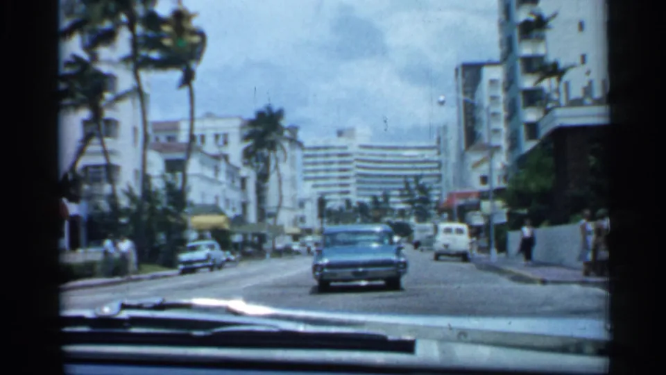 1958 Florida driving view with classic cars, palm trees, pedestrians, windshield wipers, and multi-story buildings along a broad city street.